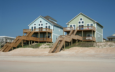 Ocean County custom home builder shows 2 homes built on dunes, the left house is blue and the house on the right is green. Both have extensive wood decking and stairs leading to beach.